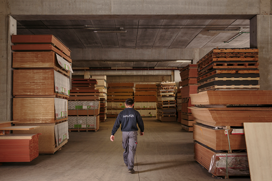 La imagen muestra el interior de un almacén industrial con grandes paneles de madera apilados a ambos lados. En el centro, una persona camina por el pasillo principal, rodeada de material de carpintería y estructuras de hormigón.