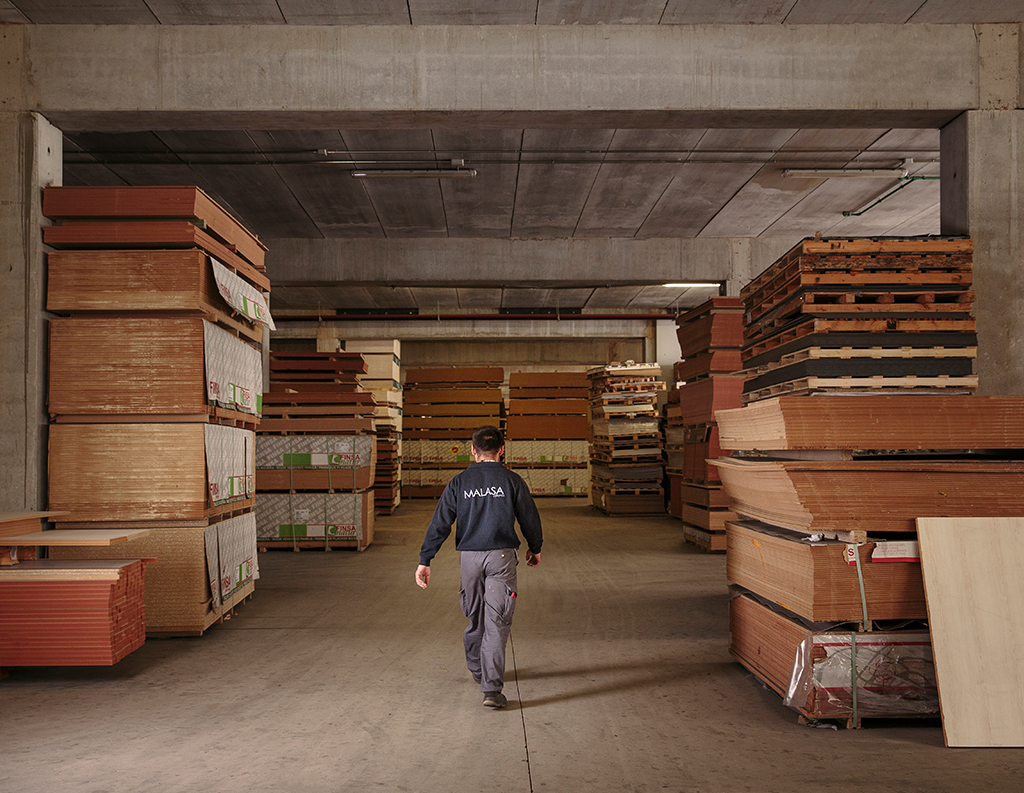 La imagen muestra el interior de un almacén industrial con grandes paneles de madera apilados a ambos lados. En el centro, una persona camina por el pasillo principal, rodeada de material de carpintería y estructuras de hormigón.