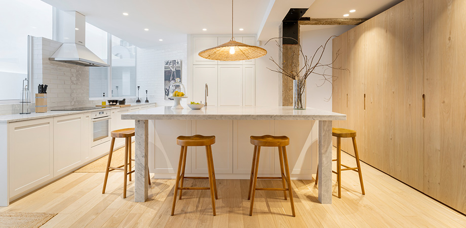 Cocina moderna de estilo minimalista con isla central de mármol, muebles blancos, taburetes de madera y acabados en madera natura