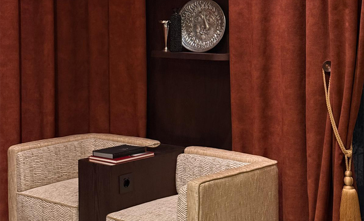 Dos sillones beige separados por un mueble oscuro ante unas cortinas rojizas elegantes.