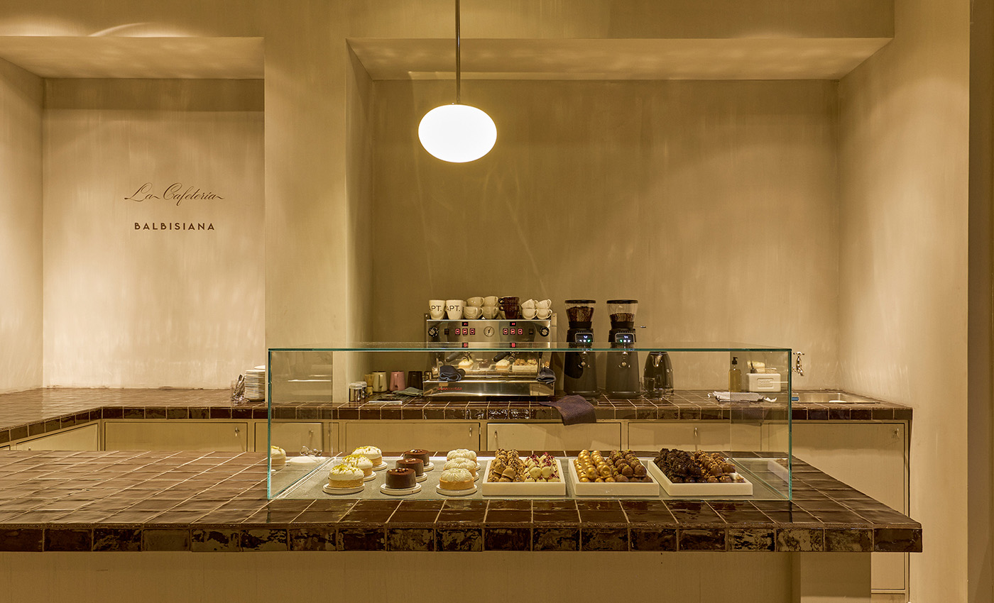 Mostrador de cafetería con vitrina llena de pasteles variados, cafetera y menaje de café en un ambiente minimalista.