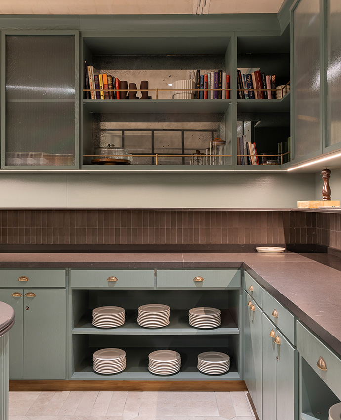 Mueble de cocina verde oliva con platos ordenados y estanterías superiores con libros.