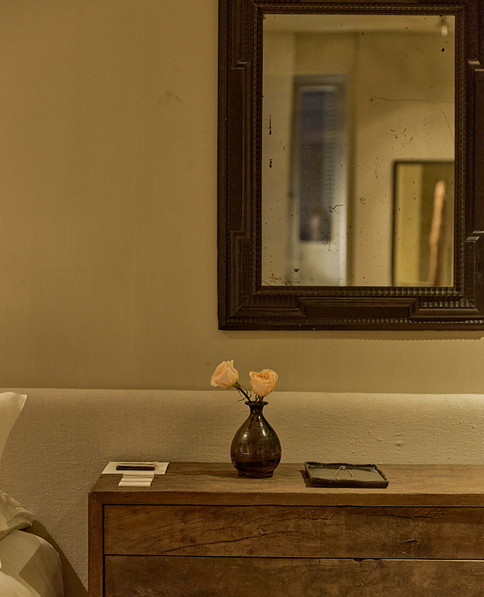 Detalle de un dormitorio con cómoda de madera rústica, jarrón con flores, un espejo antiguo y ropa de cama.