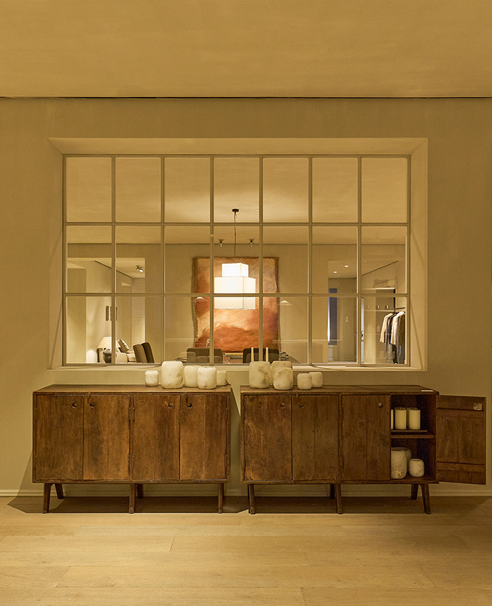 Mueble aparador bajo de madera con decoraciones, situado frente a un gran ventanal que muestra otra estancia iluminada.
