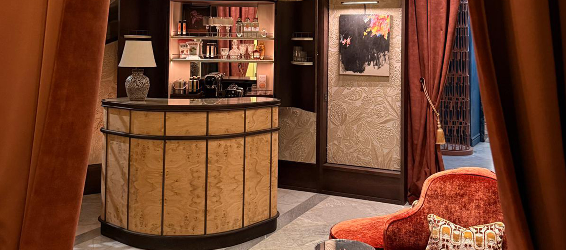 Barra de bar de madera clara con una lámpara y estantería de licores al fondo.