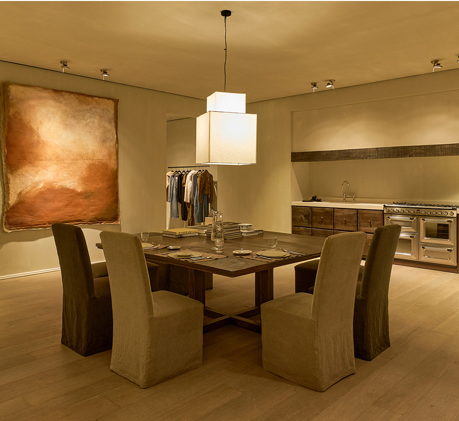 Comedor con mesa cuadrada de madera oscura, cuatro sillas tapizadas y gran cuadro abstracto, junto a una cocina moderna.