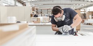 Operario con equipo de protección utiliza una lijadora orbital para pulir una pieza blanca en un taller industrial.