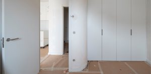 Una habitación en reformas con paredes blancas, armarios empotrados, suelo cubierto de cartón y puertas abiertas.