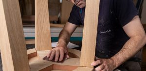Trabajador está lijando manualmente con una lija de papel la estructura de madera que es una mesa.