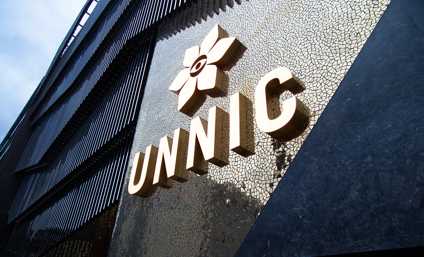 Logotipo dorado de UNNIC con una flor sobre una pared de mosaico brillante y piedra oscura texturizada.