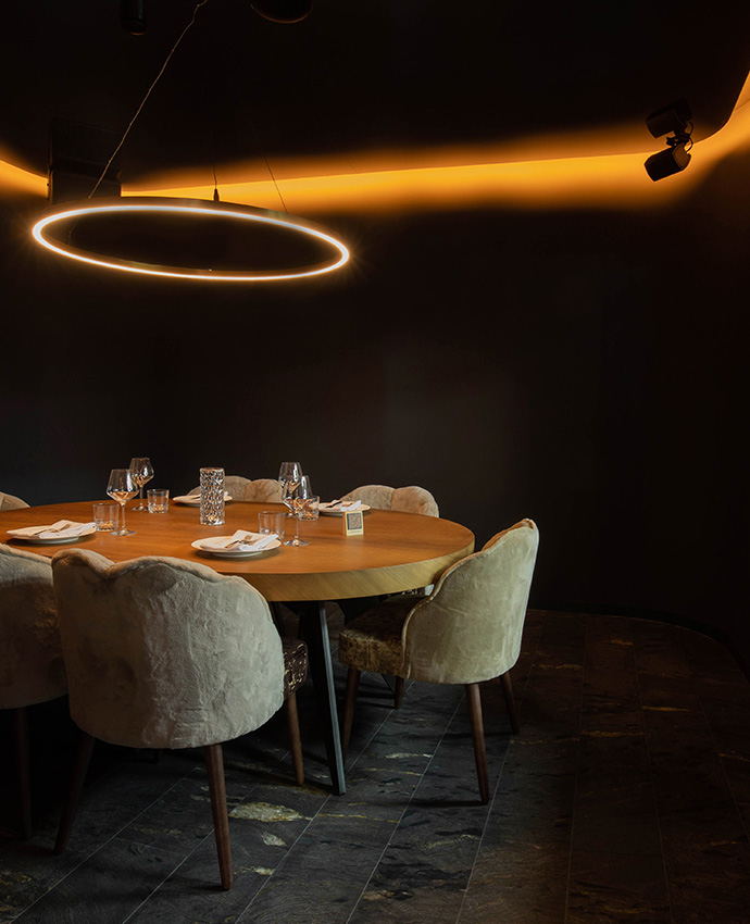 Mesa redonda de madera con sillas elegantes de terciopelo bajo una gran lámpara circular de luz cálida.