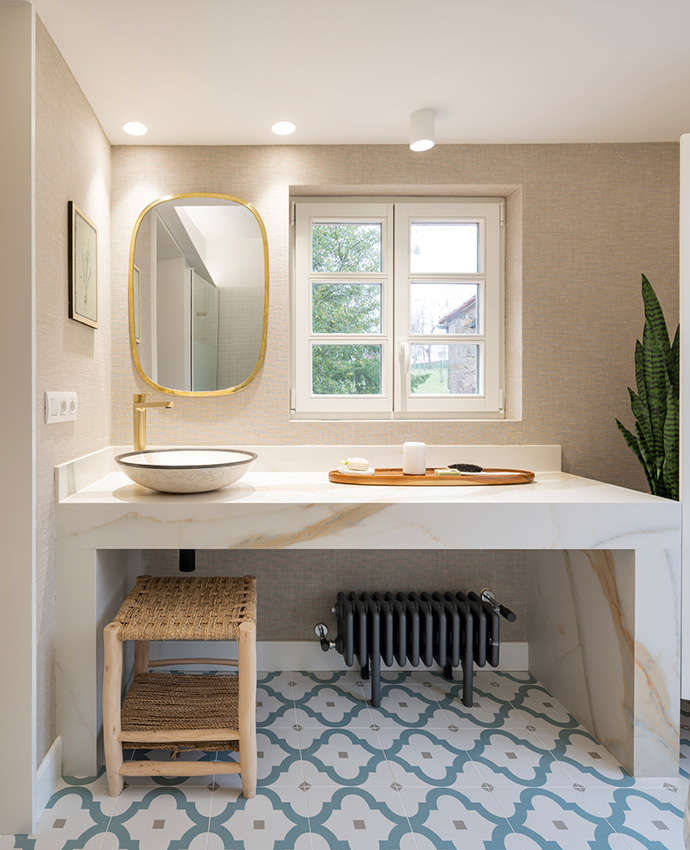 Baño con encimera de mármol, lavabo de bol, grifo dorado, espejo ovalado, suelo hidráulico azul y ventana.