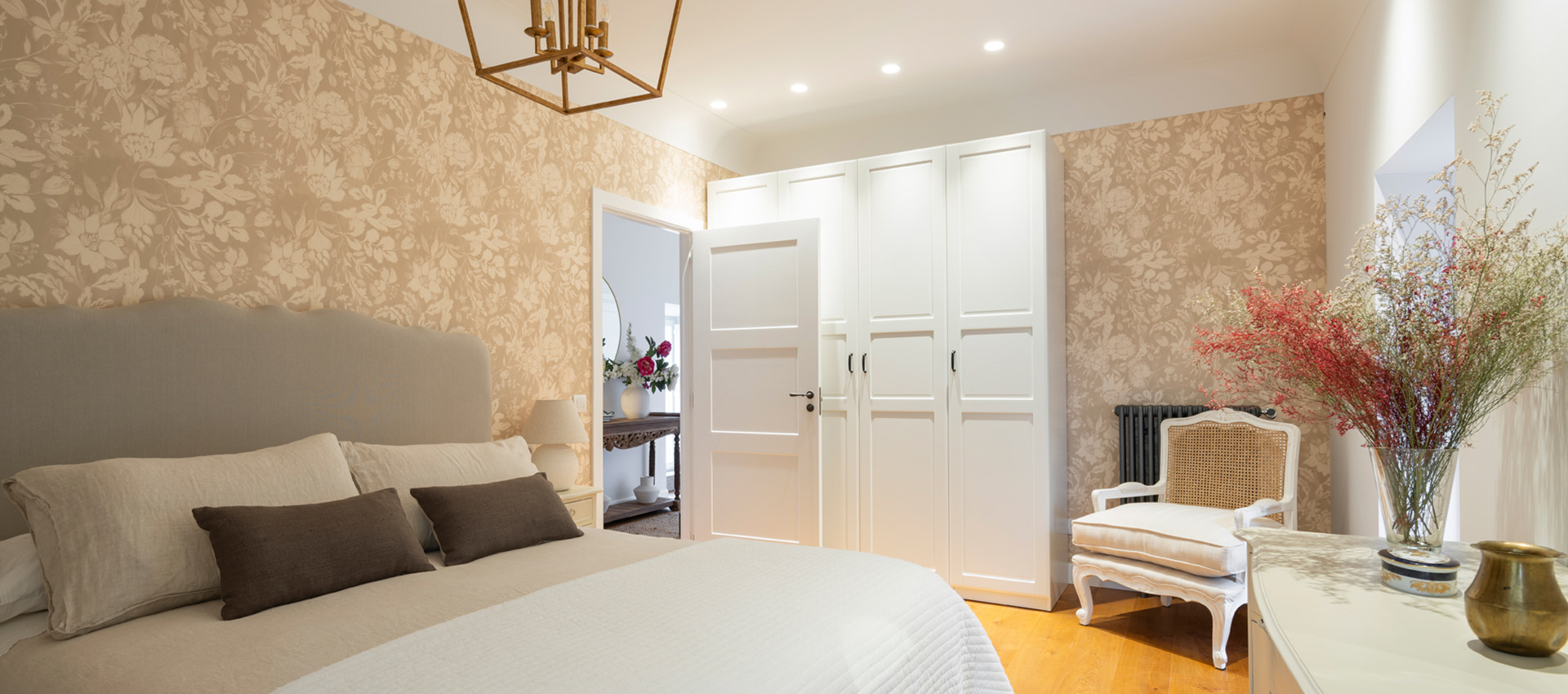 Dormitorio con papel pintado floral, cama grande, armarios blancos, silla de rejilla y jarrón con flores.