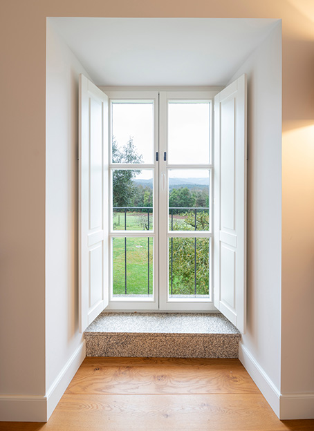 Vista de una ventana de madera blanca abierta con contraventanas interiores, mostrando un jardín verde exterior.