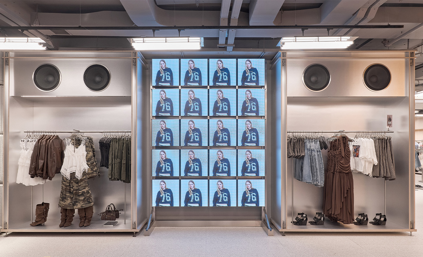 Exhibidor metálico de ropa con percheros, grandes altavoces integrados y una pantalla central cuadriculada con imágenes sincronizadas.
