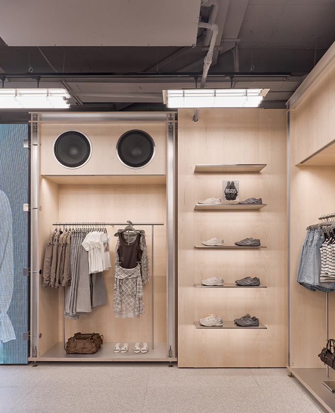 Muebles de madera en tienda con percheros de ropa casual, estanterías con zapatillas deportivas y altavoces integrados.