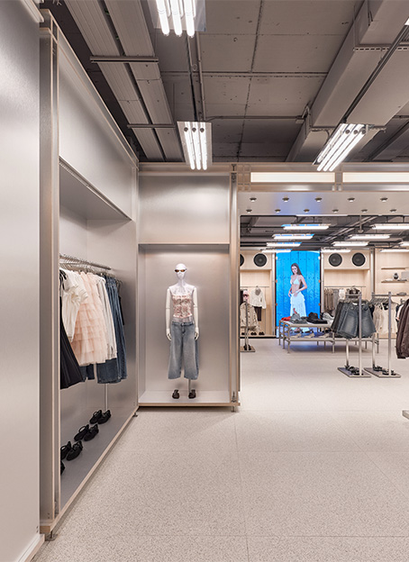 Pasillo de tienda de ropa con acabados metálicos, maniquí expuesto, luces fluorescentes y pantalla digital al fondo.