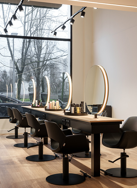 Zona de peluquería con una larga mesa de madera, espejos redondos con luz perimetral y sillones negros frente a una gran cristalera.