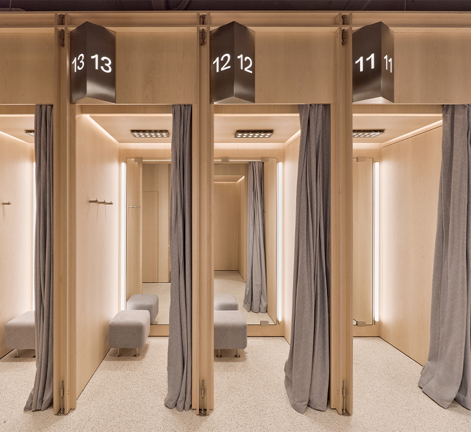 Probadores individuales de madera numerados con carteles metálicos, cortinas grises de tela y banquetas tapizadas en su interior.