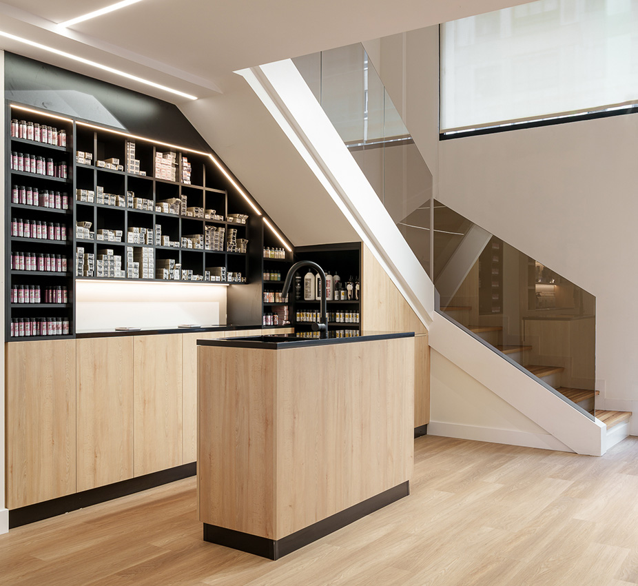 Perspectiva interior que muestra un mueble negro con estantes para productos, una barra con lavabo y la escalera de cristal al fondo.
