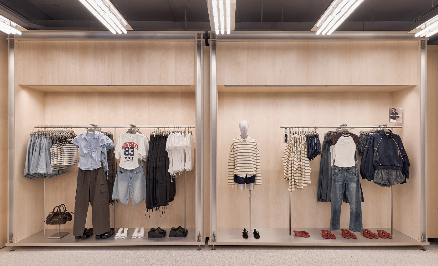 Muebles de madera clara en pared con percheros de ropa variada, maniquí central vestida y zapatos organizados abajo.