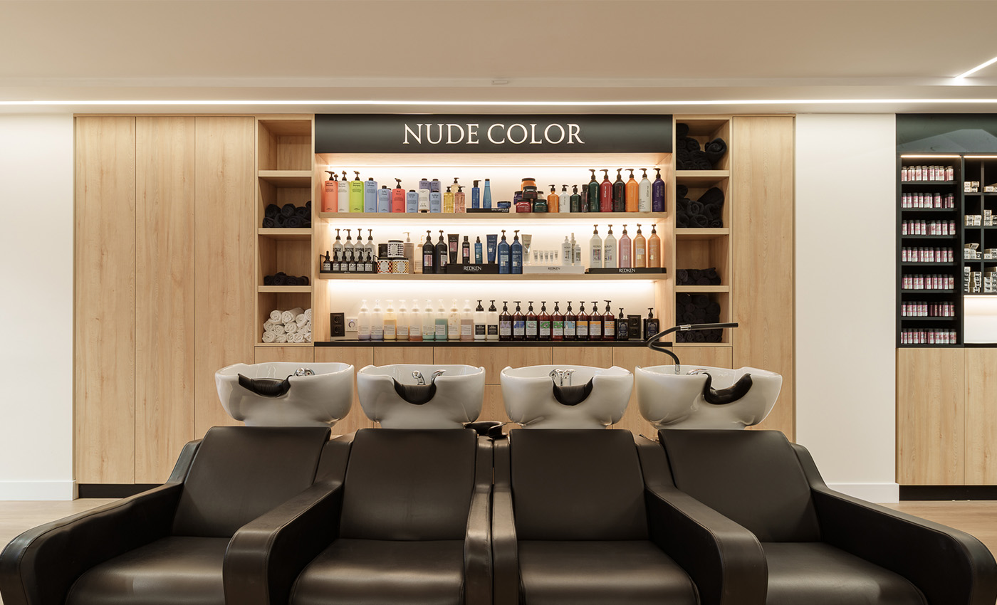 Área de lavado de cabello con cuatro sillones negros orientados hacia un gran expositor de madera iluminado con el cartel Nude Color.