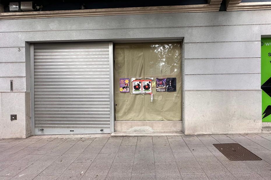 Fachada exterior de un local comercial cerrado con persiana metálica y varios carteles pegados en el escaparate.