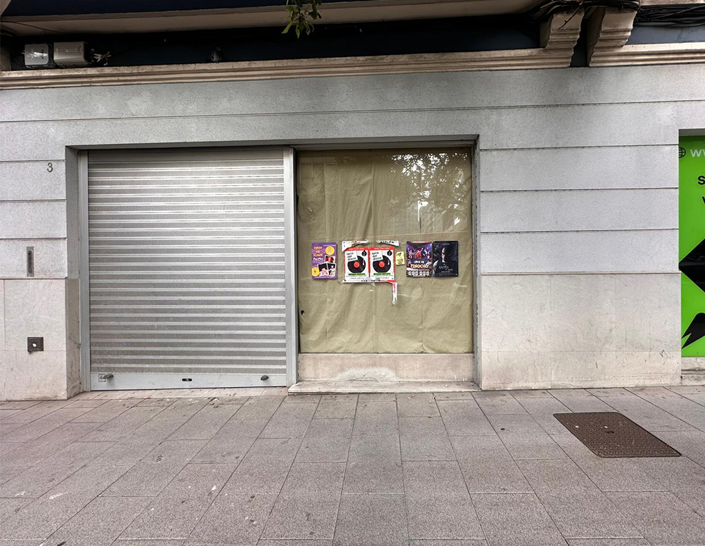 Fachada exterior de un local comercial cerrado con persiana metálica y varios carteles pegados en el escaparate.