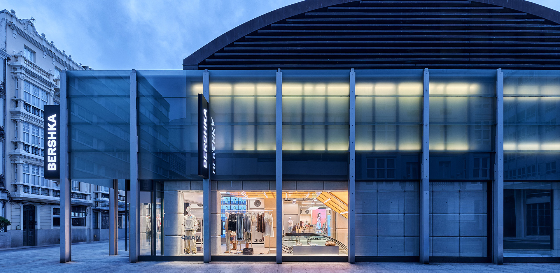 Vista frontal nocturna de tienda Bershka con rótulos luminosos, fachada de vidrio y amplio escaparate al público.