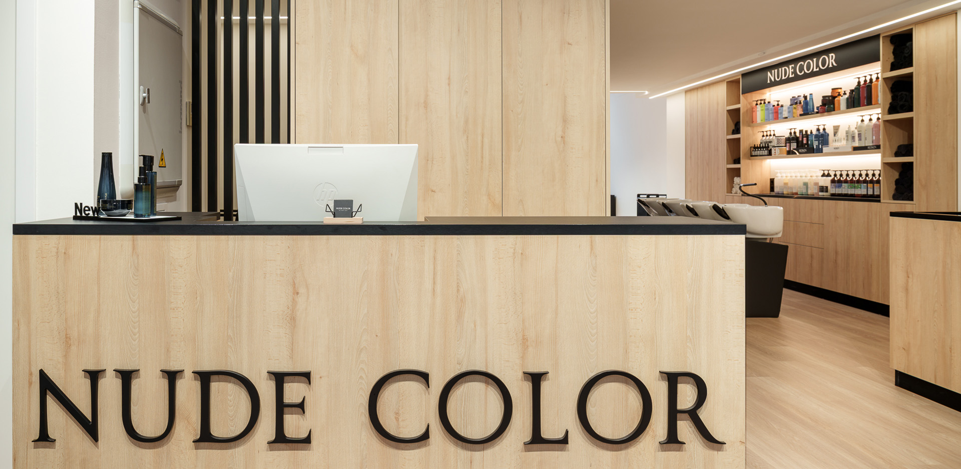 Mostrador de recepción de madera con el logotipo Nude Color en grandes letras oscuras y un ordenador blanco encima.