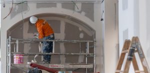 Obrero equipado con casco y chaqueta naranja realizando trabajos de enlucido sobre un andamio en la obra.