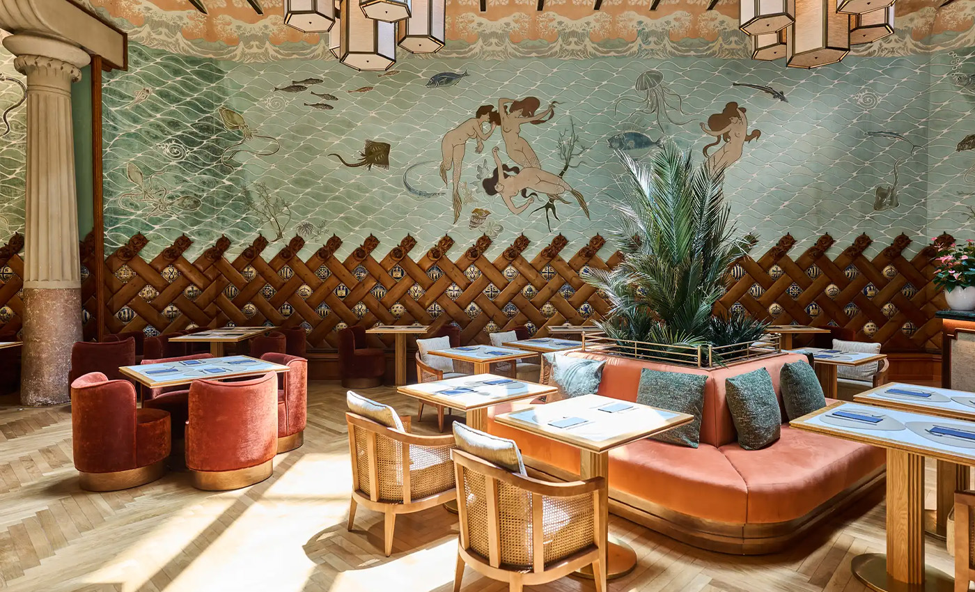 Salón de restaurante decorado con murales marinos, sofás curvos, plantas y mesas elegantes iluminadas.