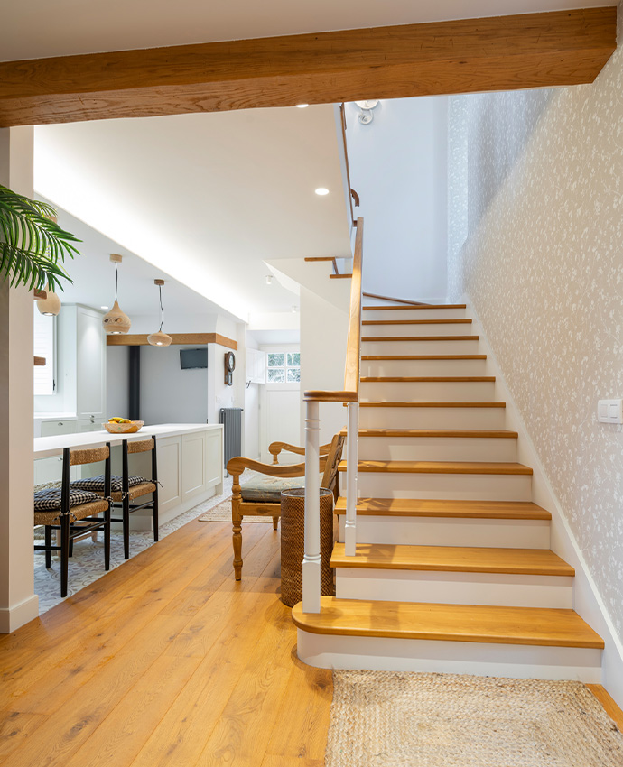 Interior luminoso con escalera de madera y blanca, vigas expuestas, cocina abierta al fondo y suelo de parqué.
