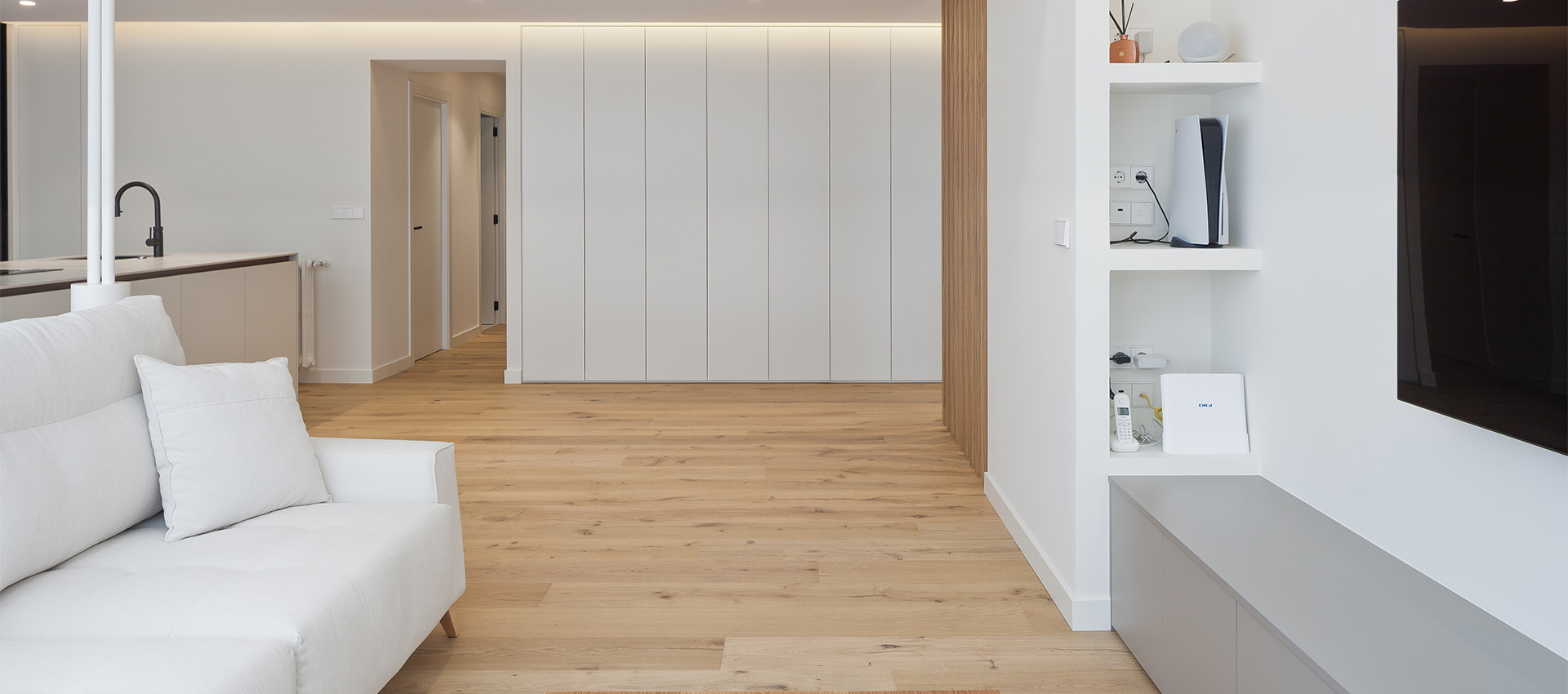 Vista del salón que muestra el sofá blanco, estanterías empotradas con dispositivos electrónicos y puertas paneladas al fondo.