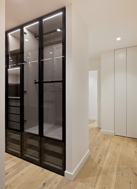 Detalle de un armario vestidor de diseño moderno con puertas de vidrio transparente y perfilería negra, situado junto a un pasillo con suelo de madera.