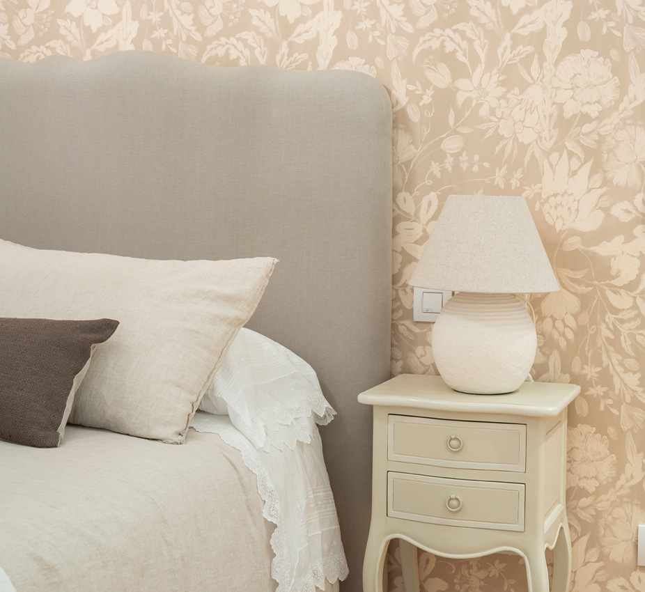 Cama con cabecero tapizado gris, cojines neutros, mesilla blanca con lámpara y papel pintado floral beige.