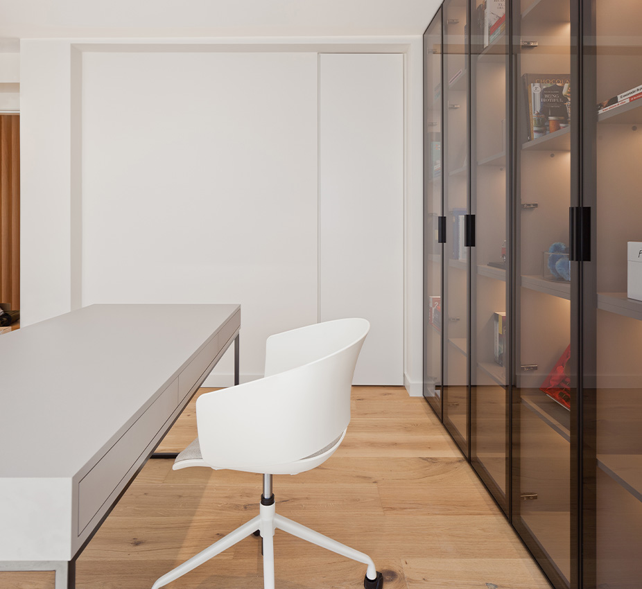 Zona de estudio o despacho con escritorio de líneas depuradas, silla ergonómica blanca y gran estantería de cristal al fondo.