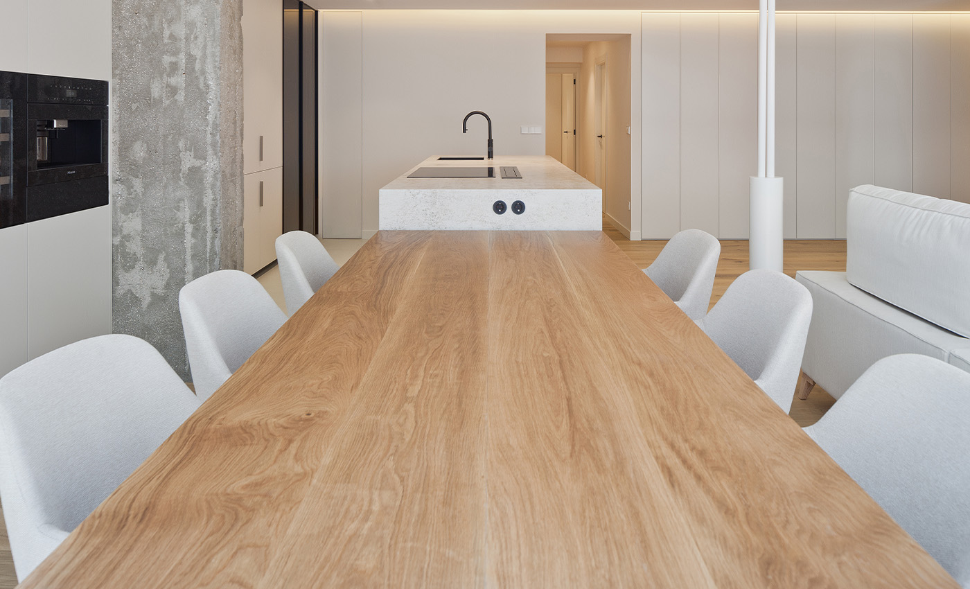 Perspectiva longitudinal de la mesa de madera del comedor conectada directamente con la isla de la cocina y una columna de hormigón visto.