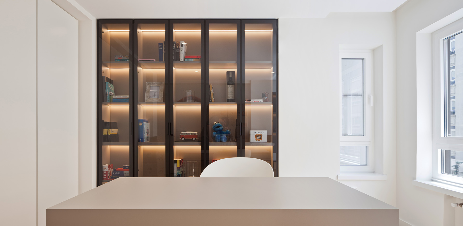 Despacho minimalista con gran librería de puertas de cristal con iluminación LED interior, escritorio claro y dos ventanas laterales.