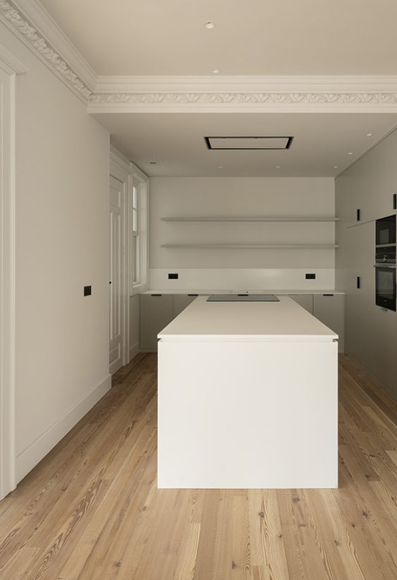 Pasillo interior minimalista con techo de hormigón visto, suelo de madera clara y cocina pequeña con isla metálica al fondo.