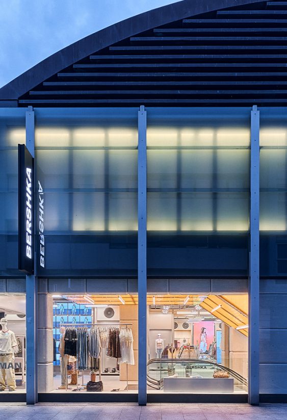 Vista frontal nocturna de tienda Bershka con rótulos luminosos, fachada de vidrio y amplio escaparate al público.