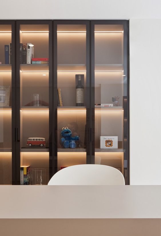 Despacho minimalista con gran librería de puertas de cristal con iluminación LED interior, escritorio claro y dos ventanas laterales.