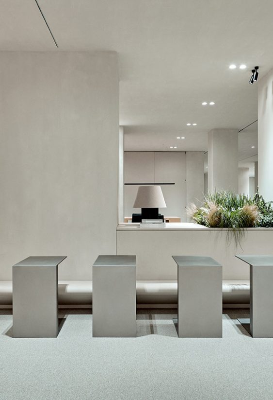 Interior minimalista de centro de trabajo con escalones de madera a la izquierda, cinco pedestales metálicos en primer plano y jardinera con plantas.