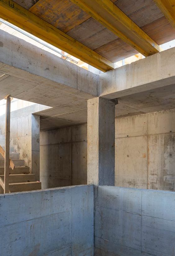 Interior de vivienda en construcción con estructura de hormigón, escalera provisional y vigas de madera en el techo.