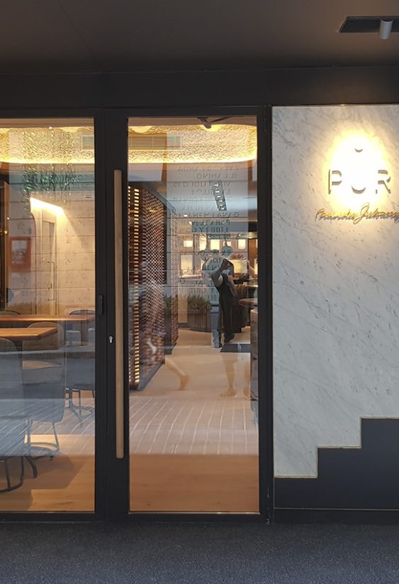 Fachada del cocktail bar Impür con logo iluminado y vista del interior a través de puertas de vidrio.