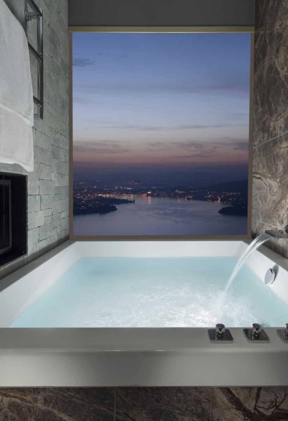 Baño de spa con bañera empotrada, chimenea integrada en muro de piedra y ventana con vista panorámica nocturna.